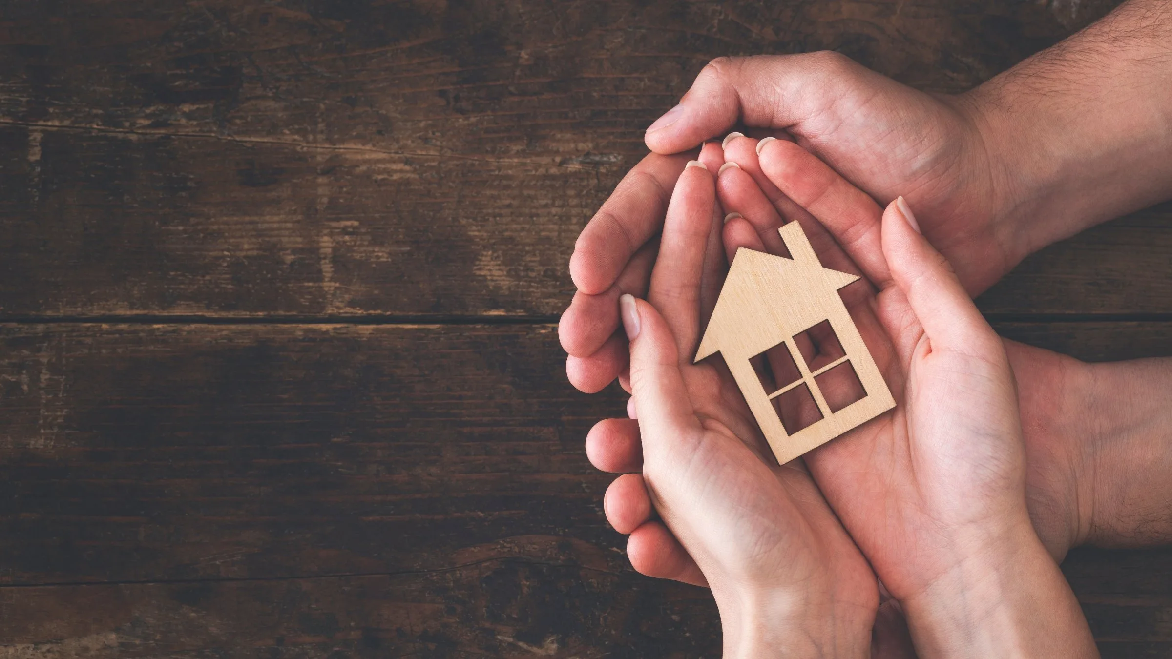 Hands gently cradle a small wooden house model on a dark wooden surface, symbolizing home security and care.