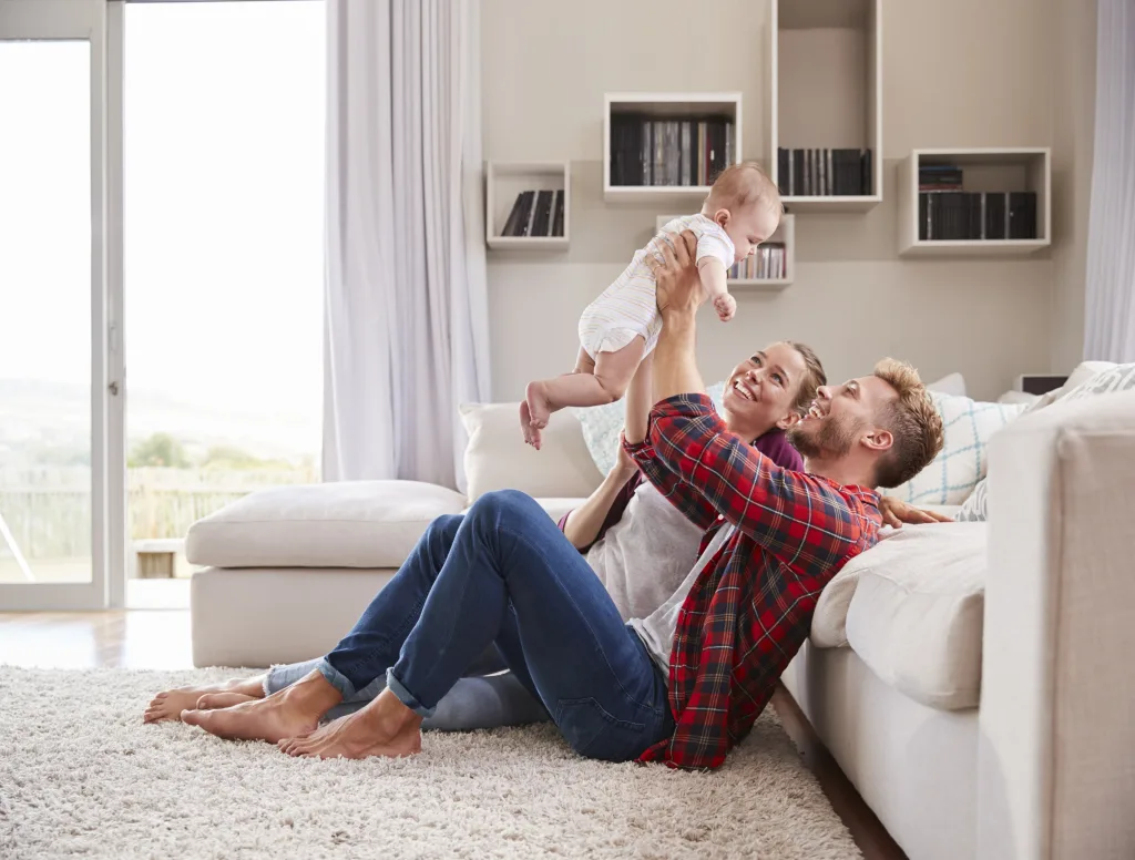 Happy parents sitting on the living room rug lifting their baby playfully in the air.