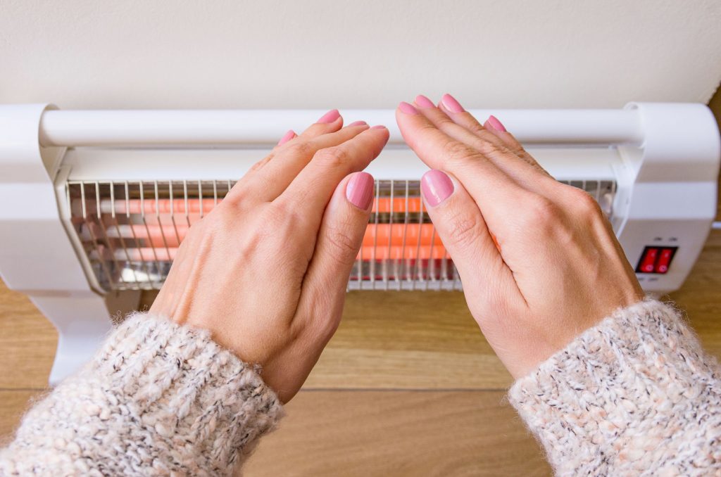 Close-up of a person warming their hands over a glowing electric space heater.