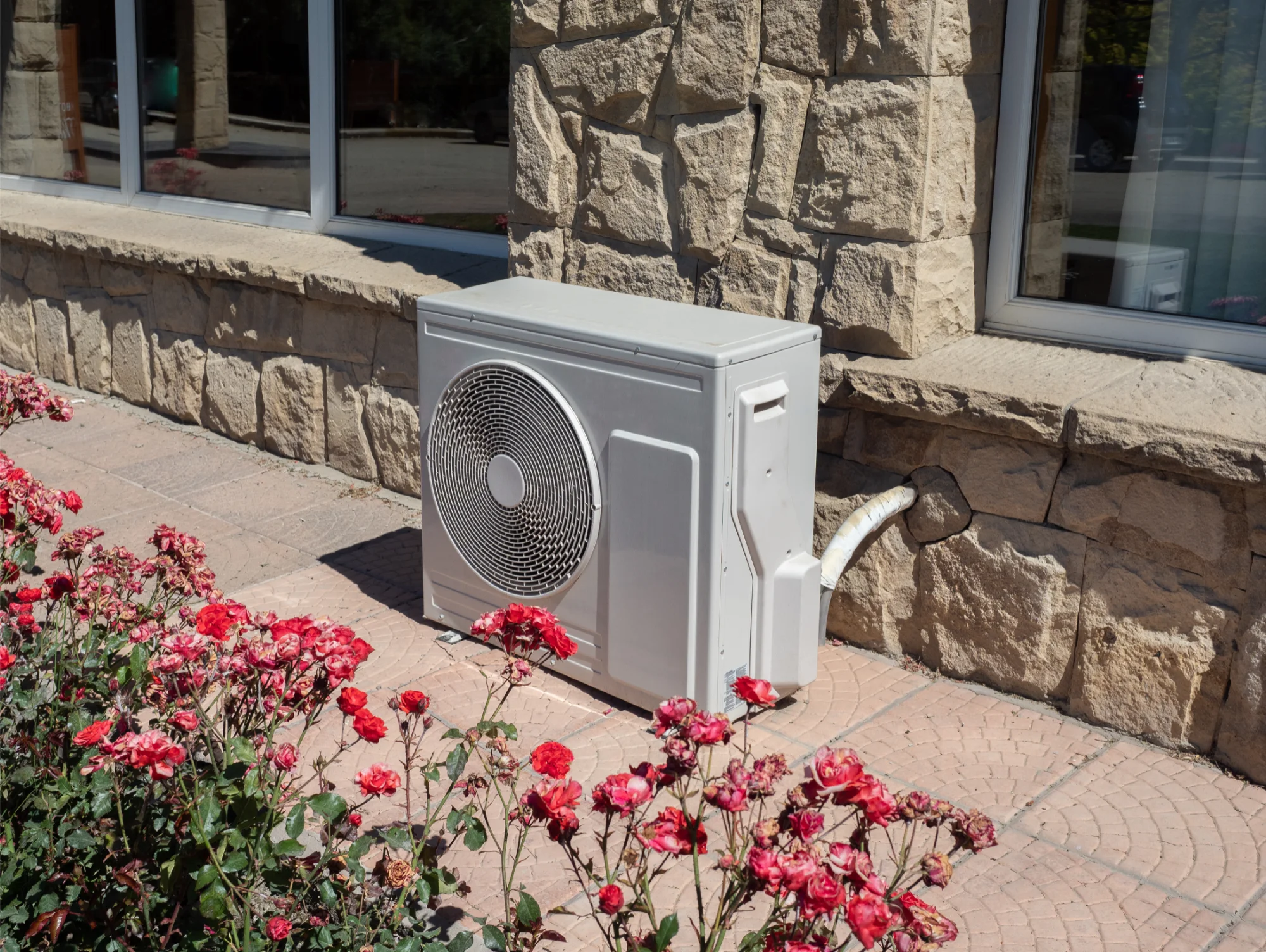 Outdoor air conditioning unit against a stone wall, surrounded by blooming red flowers on a sunny day. The scene conveys a sense of warmth and serenity.