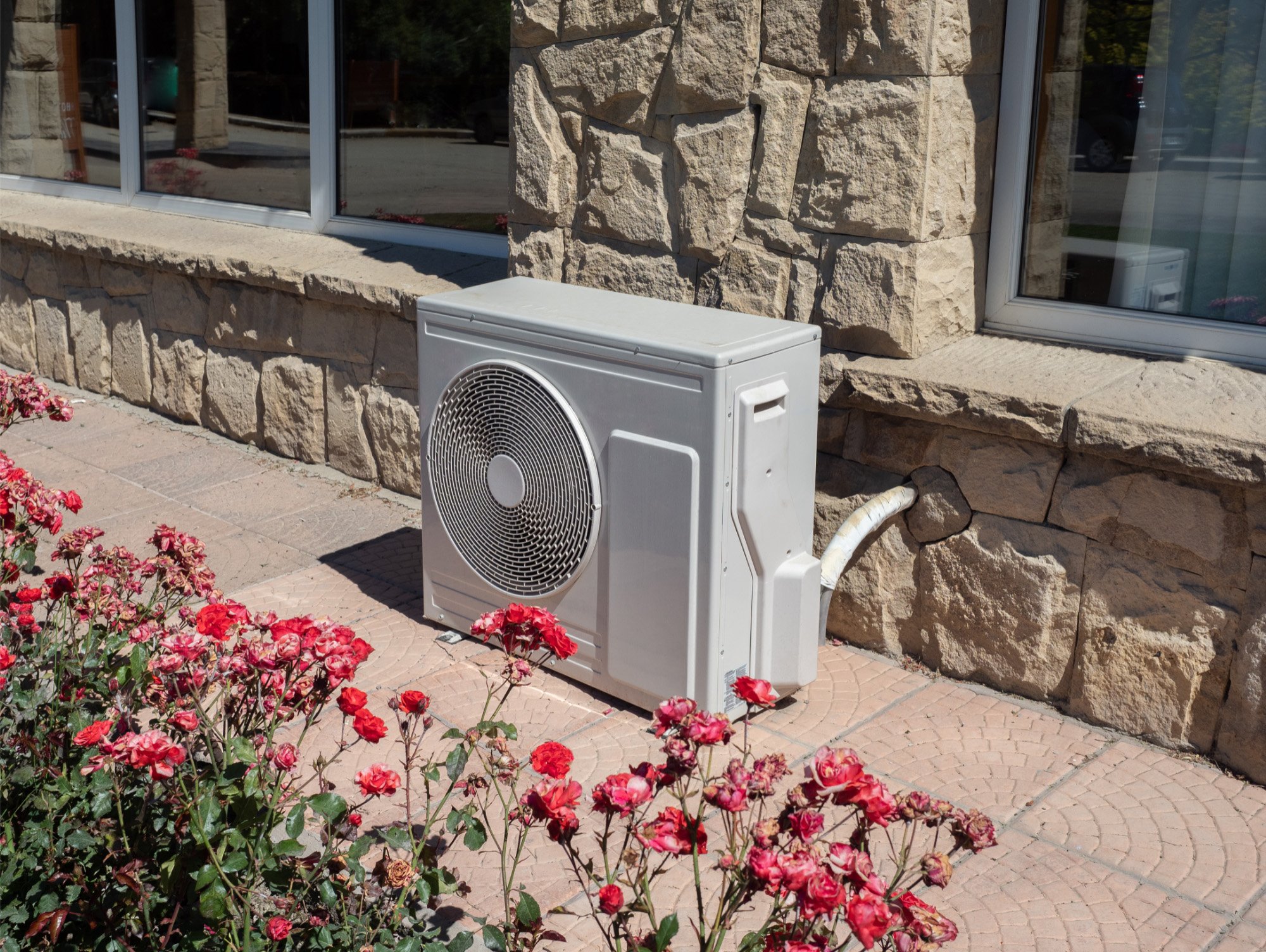 Outdoor air conditioning unit against a stone wall, surrounded by blooming red flowers on a sunny day. The scene conveys a sense of warmth and serenity.