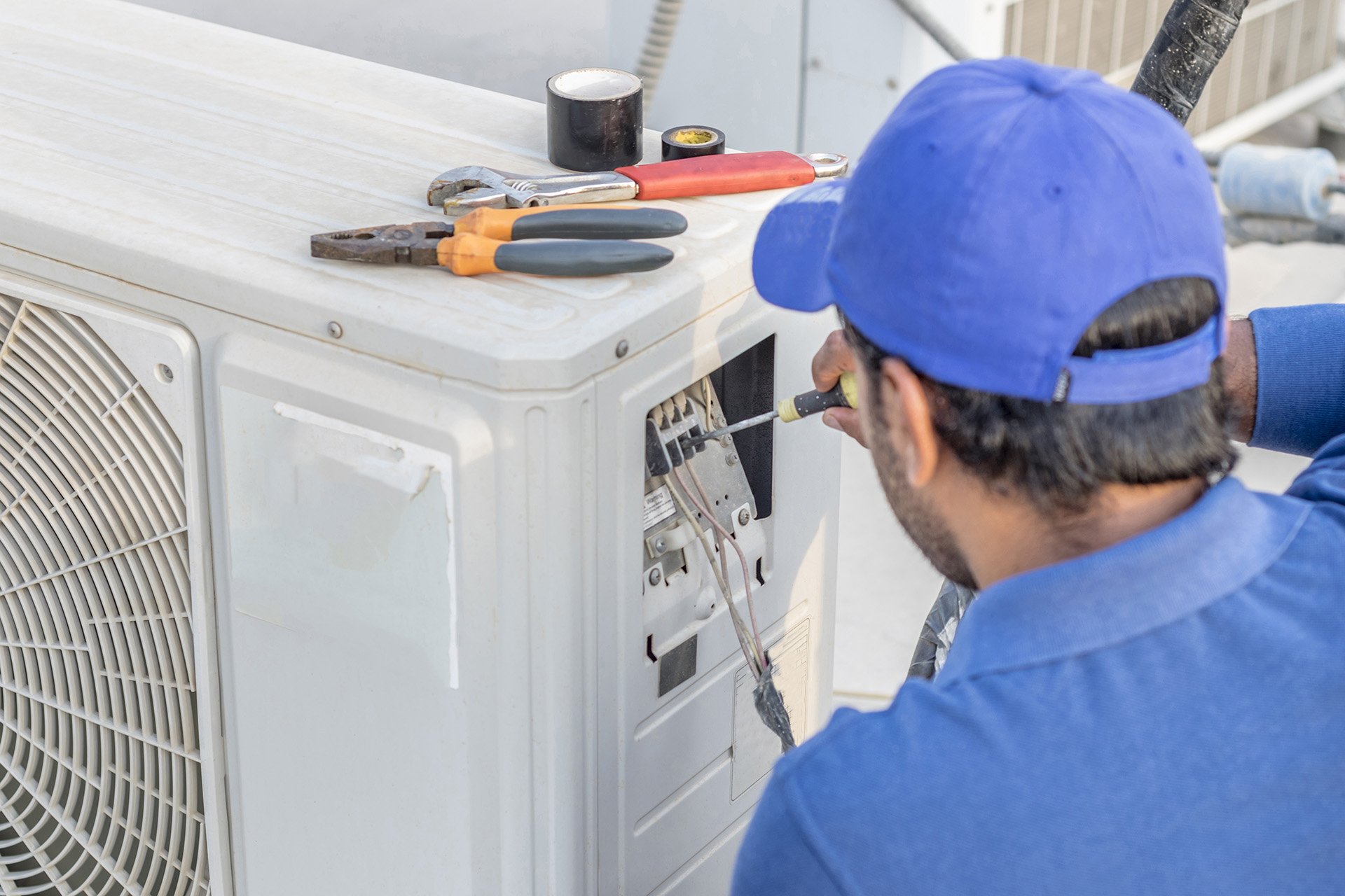 A professional electrician in a grey uniform and white cap using tools to perform AC repair in Elk Grove, CA, while servicing a heavy-duty central air conditioning unit on a rooftop.