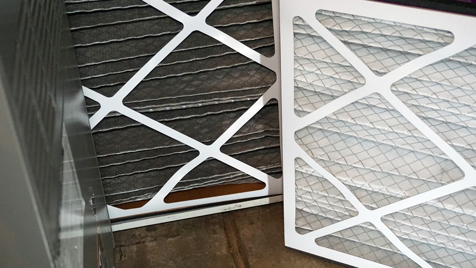 A clean air filter is leaning against a used, darker one inside an HVAC system. The contrast highlights the dirty filter's grime. The tone is utilitarian.