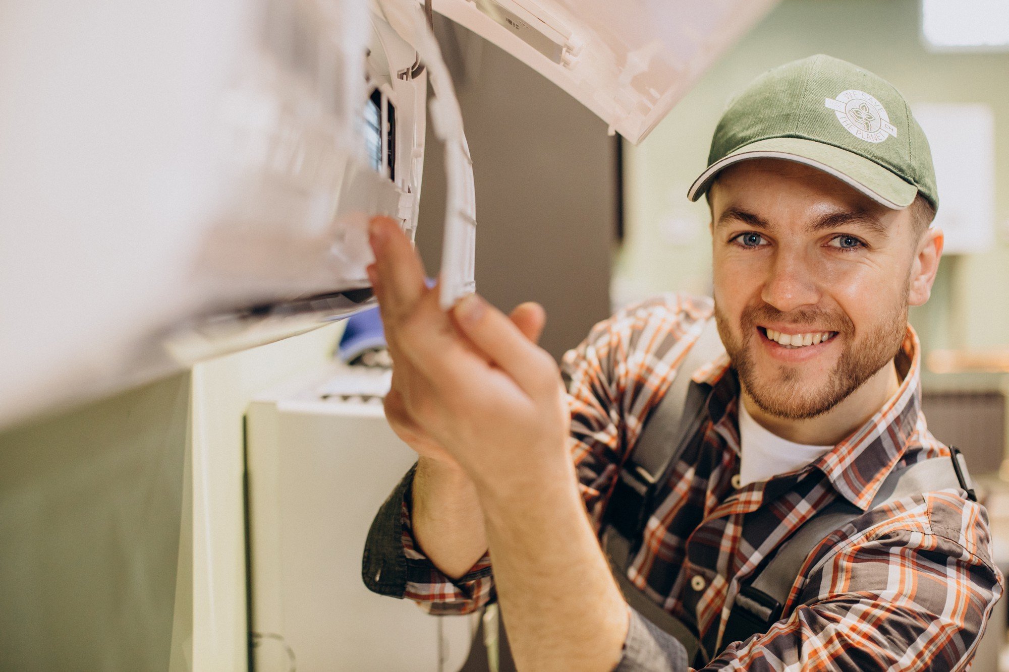 Smiling HVAC technician in a green cap and plaid shirt servicing an indoor mini-split air conditioning unit.