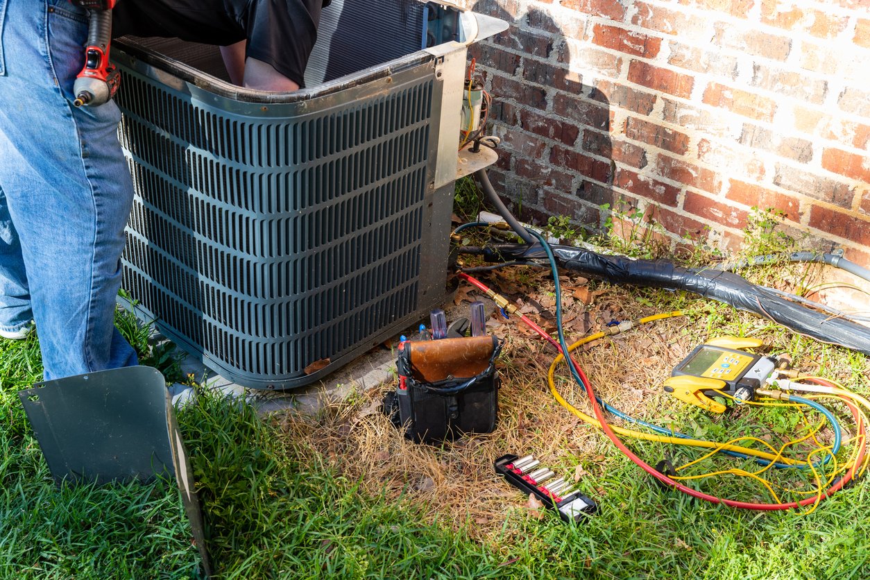 Technician repairs an outdoor air conditioning unit beside a brick wall. Tools and multicolored cables are scattered on the grass, indicating a work in progress.