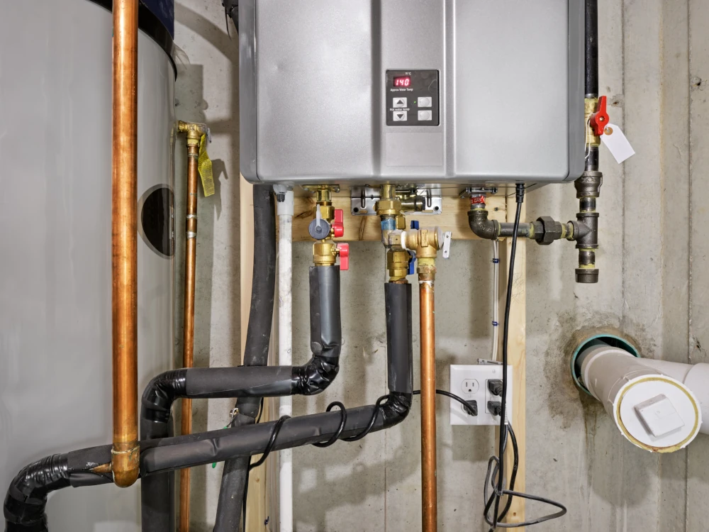 Wall-mounted boiler with digital control panel, copper pipes, insulated plumbing, valves, electrical outlet, and drain pipe in a utility room.